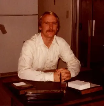 Man seated at desk, wearing white shirt.